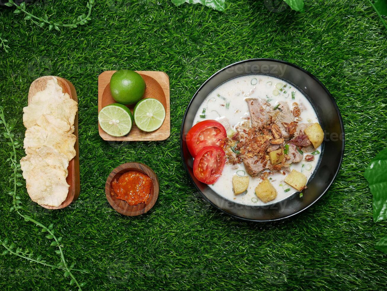 Top view Soto Betawi dish with coconut milk broth, beef, potatoes, and fried shallots. Served with fresh tomato slices, spicy sambal, crispy crackers, and fresh limes. With synthetic green grass base photo