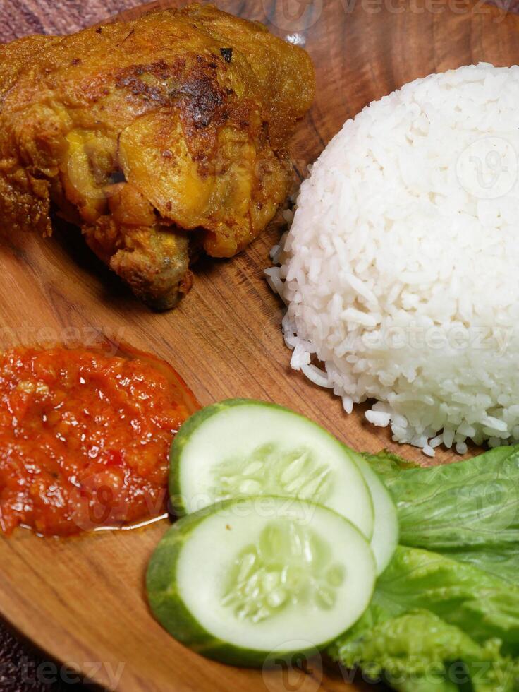 Closeup traditional Indonesian food consists of white rice, fried chicken, spicy red Sambal, fresh cucumber slices, and lettuce leaves on a wooden plate.. photo