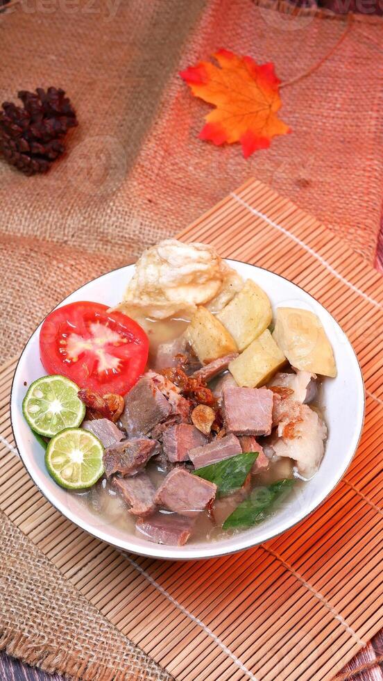Close-up of a bowl of clear beef soup with chunks of beef, potatoes, fresh tomatoes, lime slices, and fried onions. Served on bamboo mats, burlap, and leaves. photo