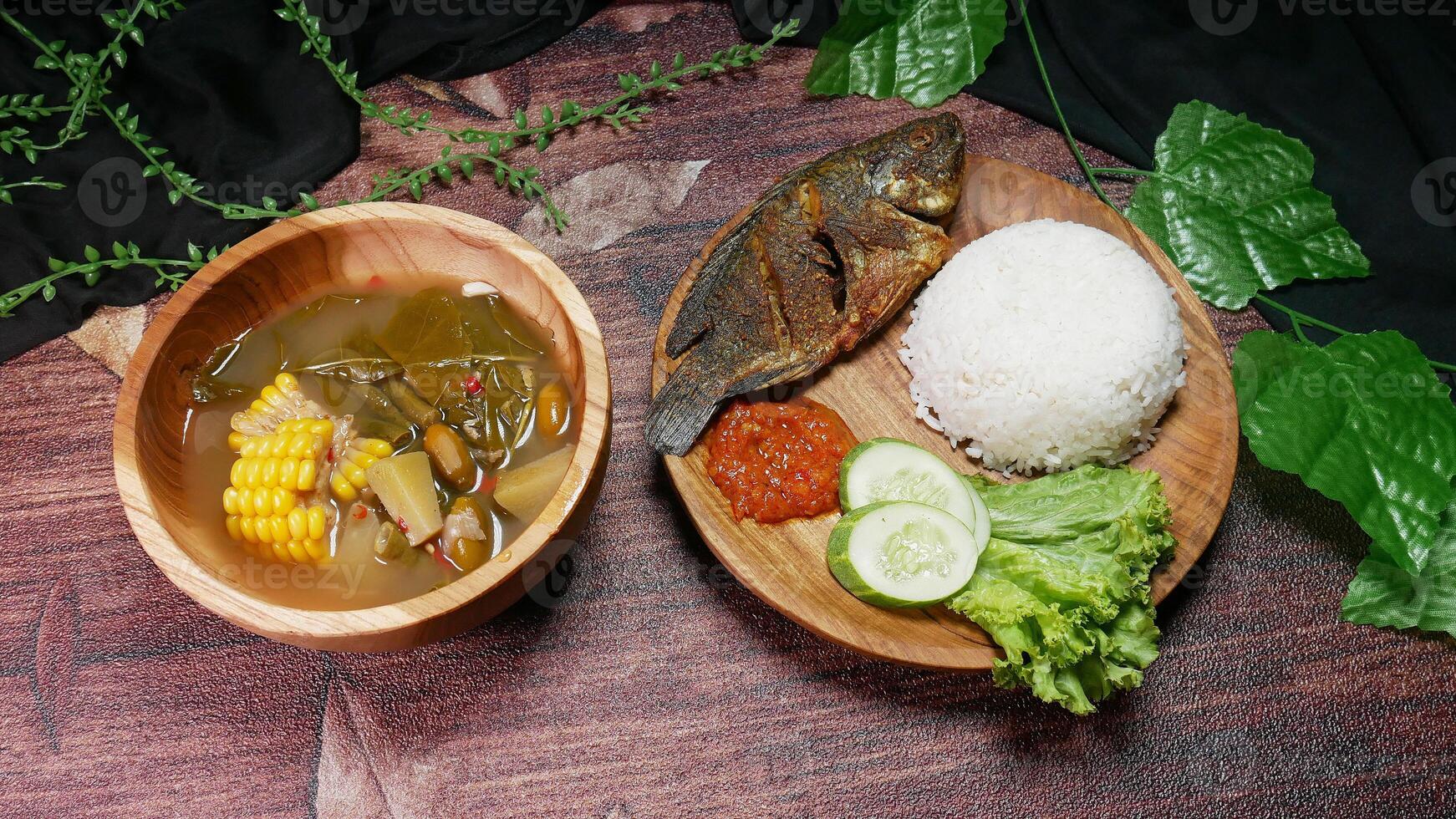 Complete traditional Indonesian food featuring fried fish, white rice, red sambal, cucumber slices, and fresh lettuce. Sayur asam in a wooden bowl with corn, chayote, long beans, and melinjo seeds. photo