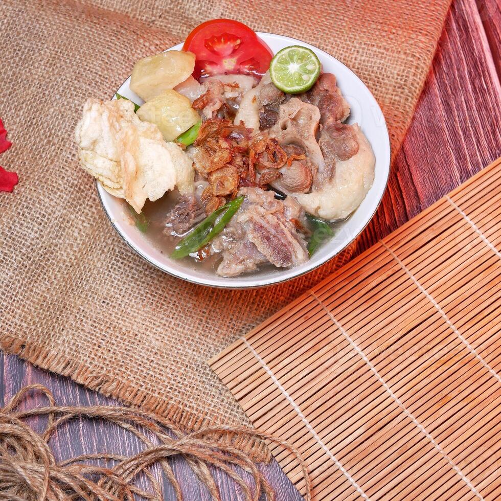 Top view of a bowl of oxtail soup or sop buntut with chunks of beef, potatoes, fresh tomatoes, lime slices, and fried onions. Served on bamboo mats, burlap, and leaves. Space for text available. photo