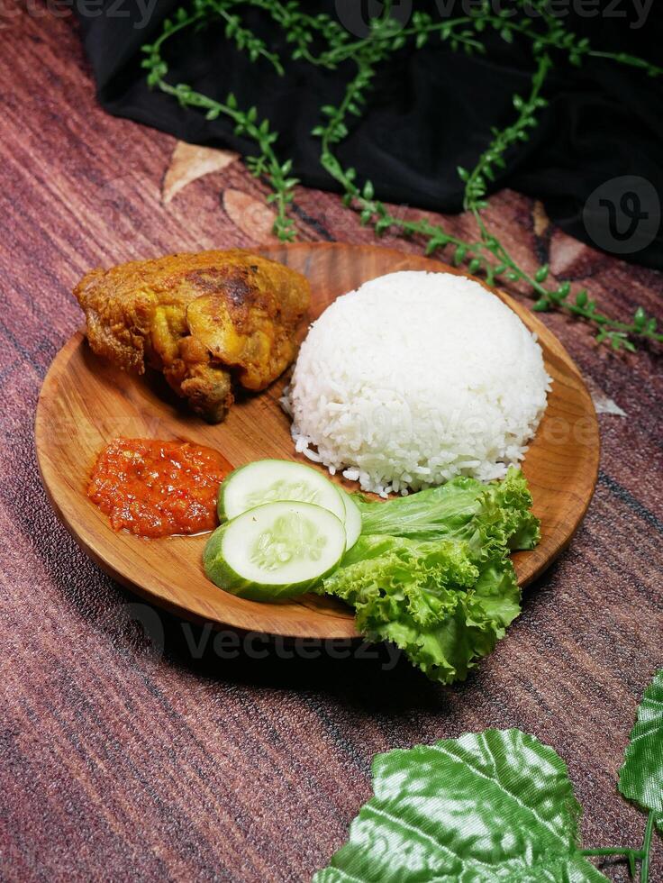 Traditional Indonesian food consists of white rice, fried chicken, spicy red Sambal, fresh cucumber slices, and lettuce leaves on a round wooden plate. Decorated with green plastic leaf. photo