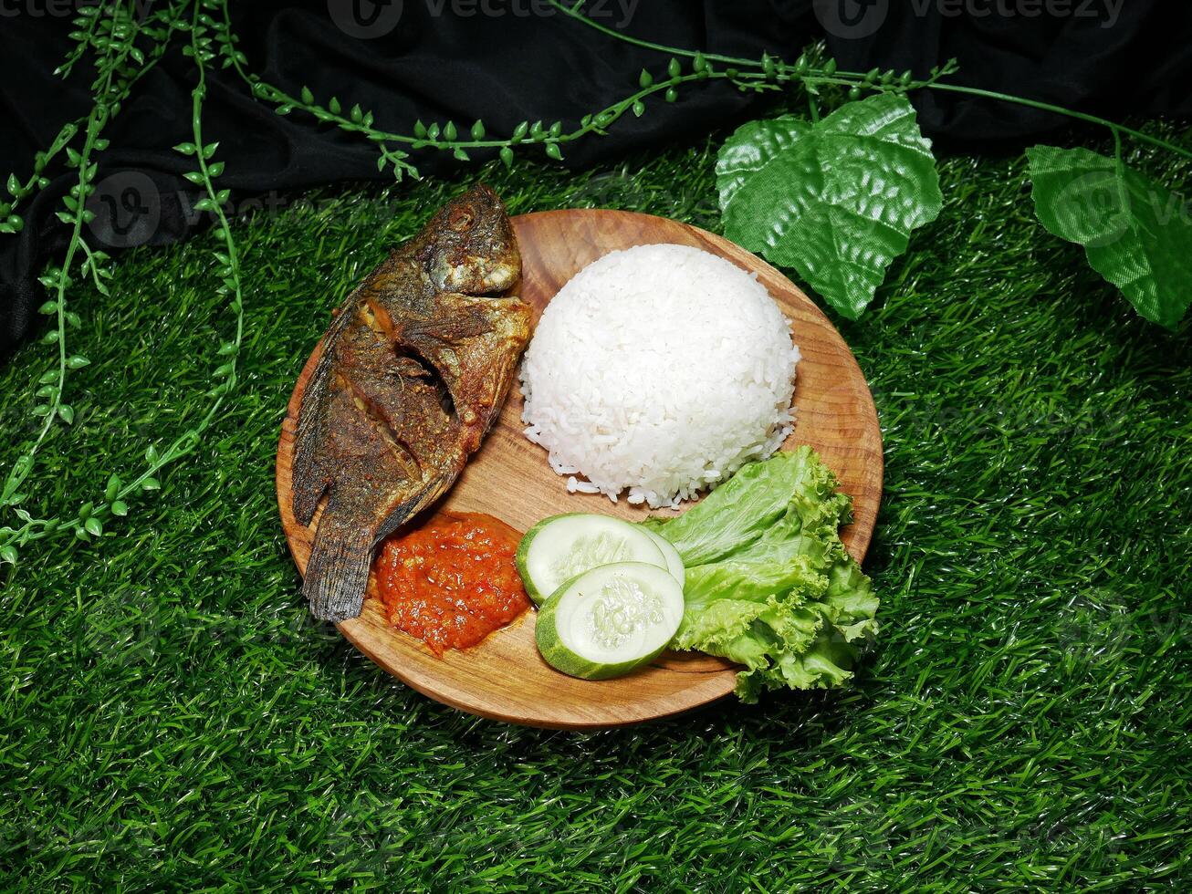 Traditional Indonesian food white rice, fried fish, spicy red Sambal, fresh cucumber slices, and lettuce, served on wooden plate. Decorated plastic leaves and with a green synthetic grass base photo