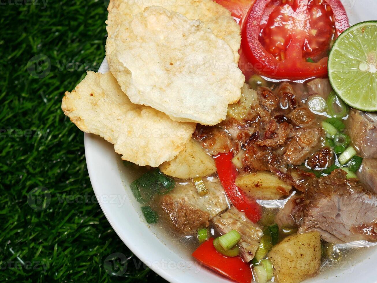 Top view close up Indonesian Soup with clear broth with Beef slices, fried Potatoes, red Tomatoes, Crackers, and Vegetables. Served in a white bowl on green synthetic grass photo
