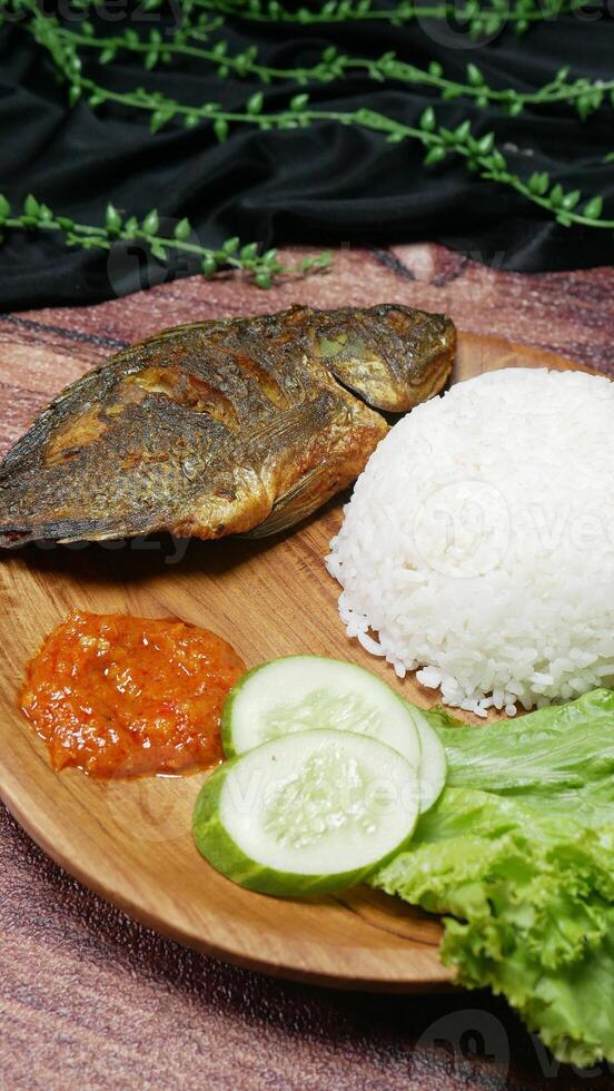 Indonesian food composition consisting of fried fish, white rice, spicy sambal, fresh cucumber slices, and green lettuce on a wooden plate with an aesthetic touch of black fabric and leaf decorations. photo