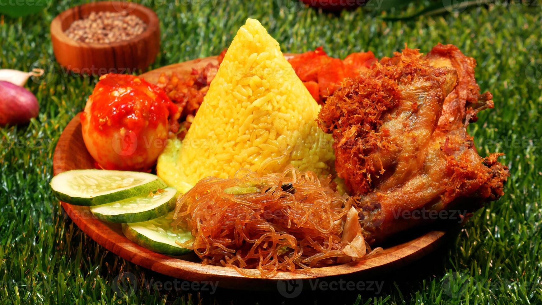 Closeup A serving of yellow rice complete with traditional Indonesian food on a wooden plate decorated with cooking spices, leaves and red flowers on a synthetic grass base. photo