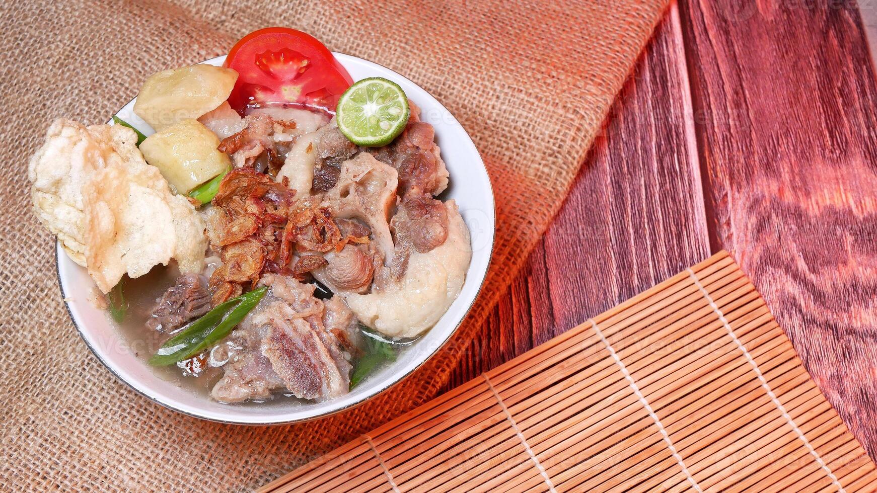 Sop buntut or oxtail soup in a white bowl with fresh potatoes, tomatoes, lime and fried onions. Served in a traditional style on a bamboo mat and burlap. Space for text available photo