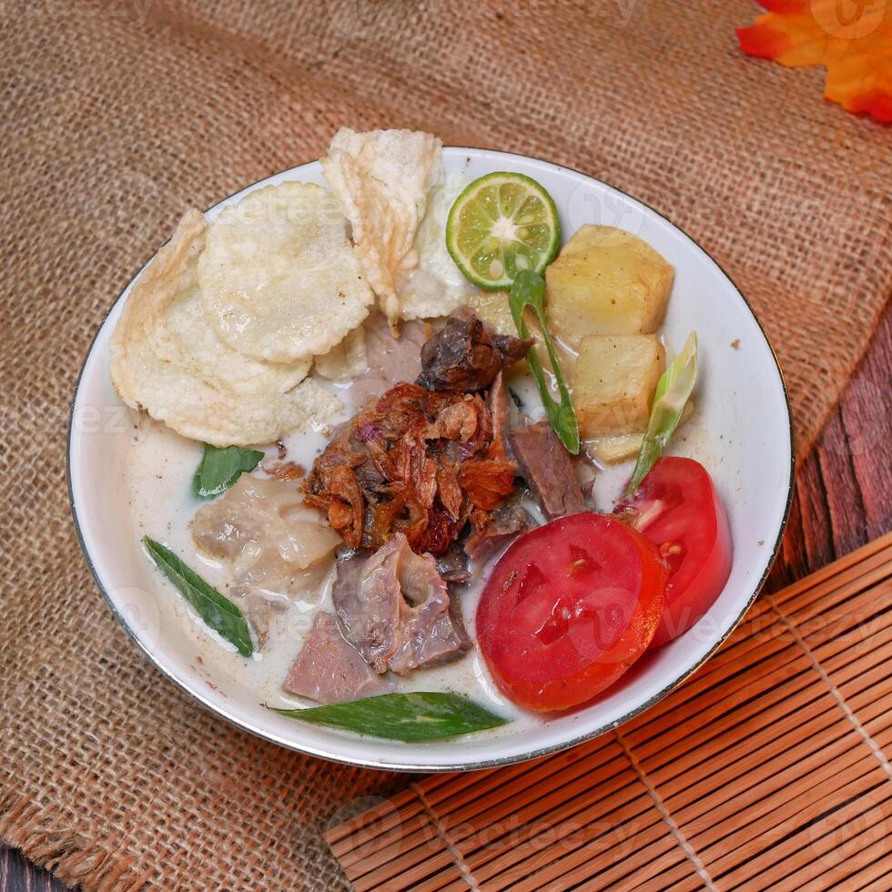 Soup or Soto Betawi with savory coconut milk broth filled with pieces of potato and beef is a traditional Indonesian food typical of Jakarta. Served in a white bowl on bamboo mat and burlap photo