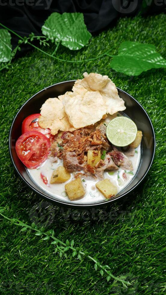 Soup or Soto Betawi with white coconut milk broth is a traditional Indonesian food typical of Jakarta. Served in a black bowl filled beef, potato, slices red tomatoes, lime, fried onions and crackers photo