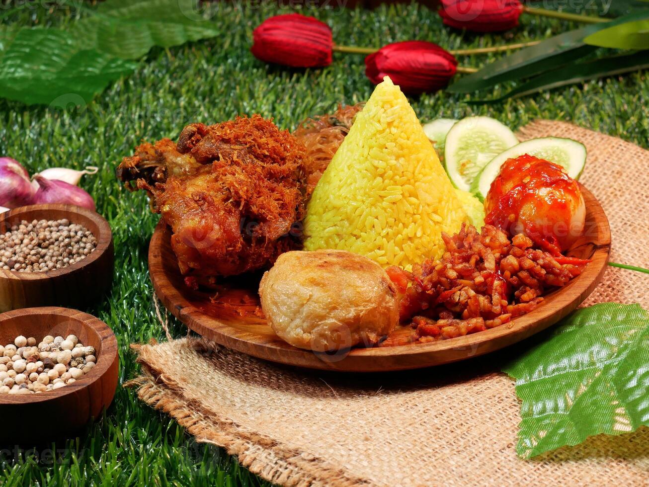 A serving of yellow rice complete with traditional Indonesian food on a wooden plate decorated with cooking spices, leaves and red flowers on a synthetic grass base. photo