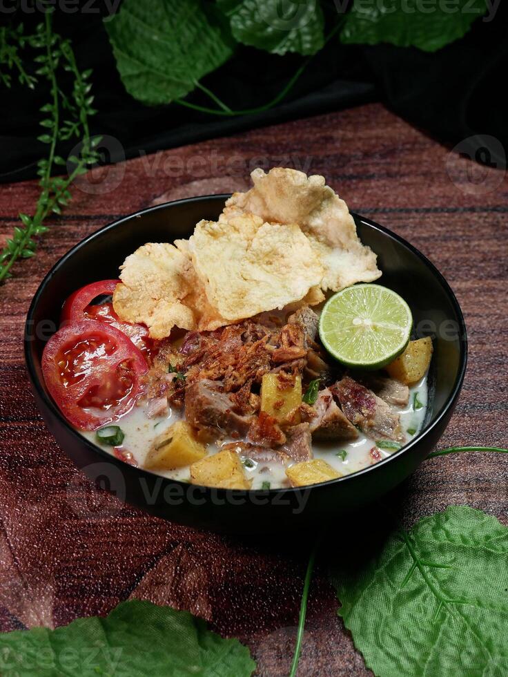 Soto Betawi with white coconut milk broth is a traditional Indonesian food typical of Jakarta. Served in a black bowl filled beef, potato, slices red tomatoes, lime, fried onions and crackers photo