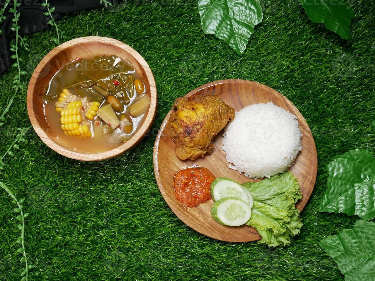 Complete traditional Indonesian food featuring fried chicken, white rice, red sambal, cucumber slices, and fresh lettuce. Sayur asam in a wooden bowl with corn, chayote, long beans, and melinjo seeds. photo