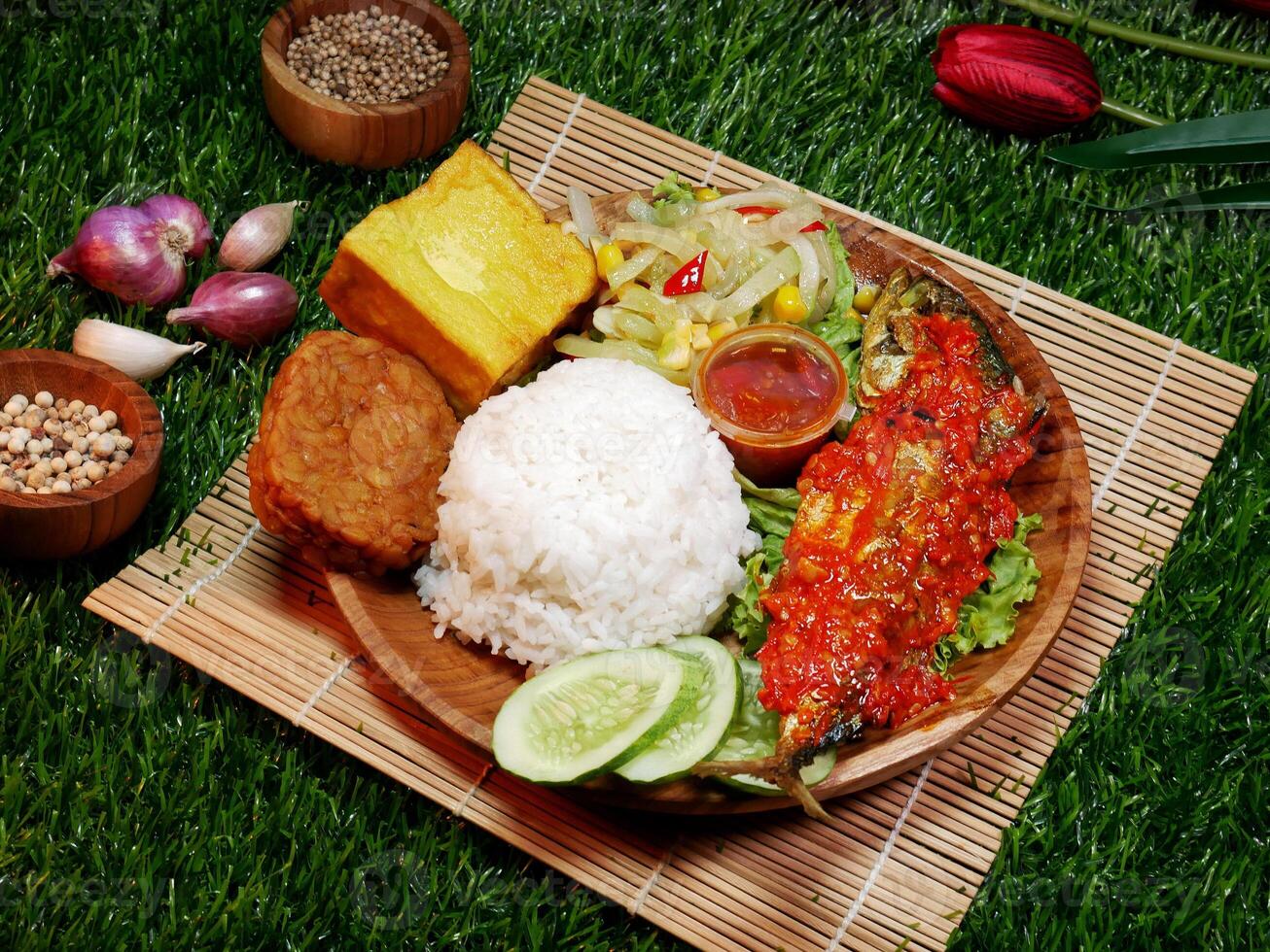 Close up A complete meal featuring white rice, fried fish with red sambal, fried tempeh, fried tofu, fresh vegetables, sambal, and cucumber slices. photo