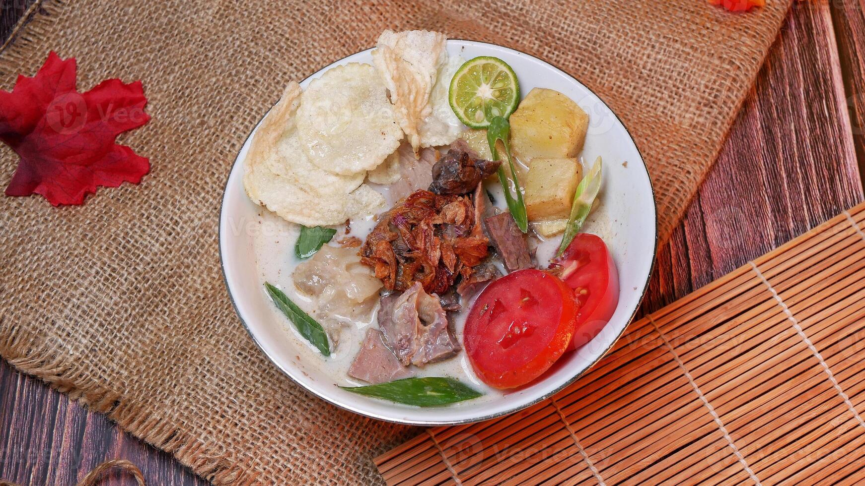 Closeup delicious Soup or Soto Betawi with beef, potato, fresh tomato, lime slices, and fried shallot. Served in white bowl with unique decoration. Space for text available photo