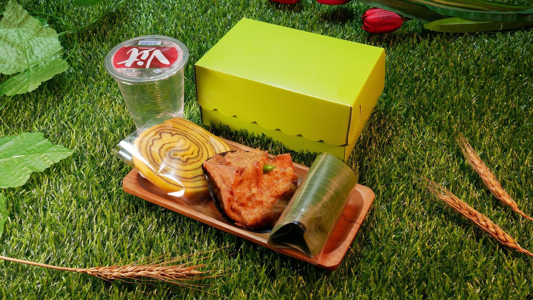 Jakarta, Indonesia. December 24, 2024. Snack box package traditional Indonesian cakes, Roll Cake, Lemper and Fried Tofu delicious and a glass of mineral water, with a natural decorative photo