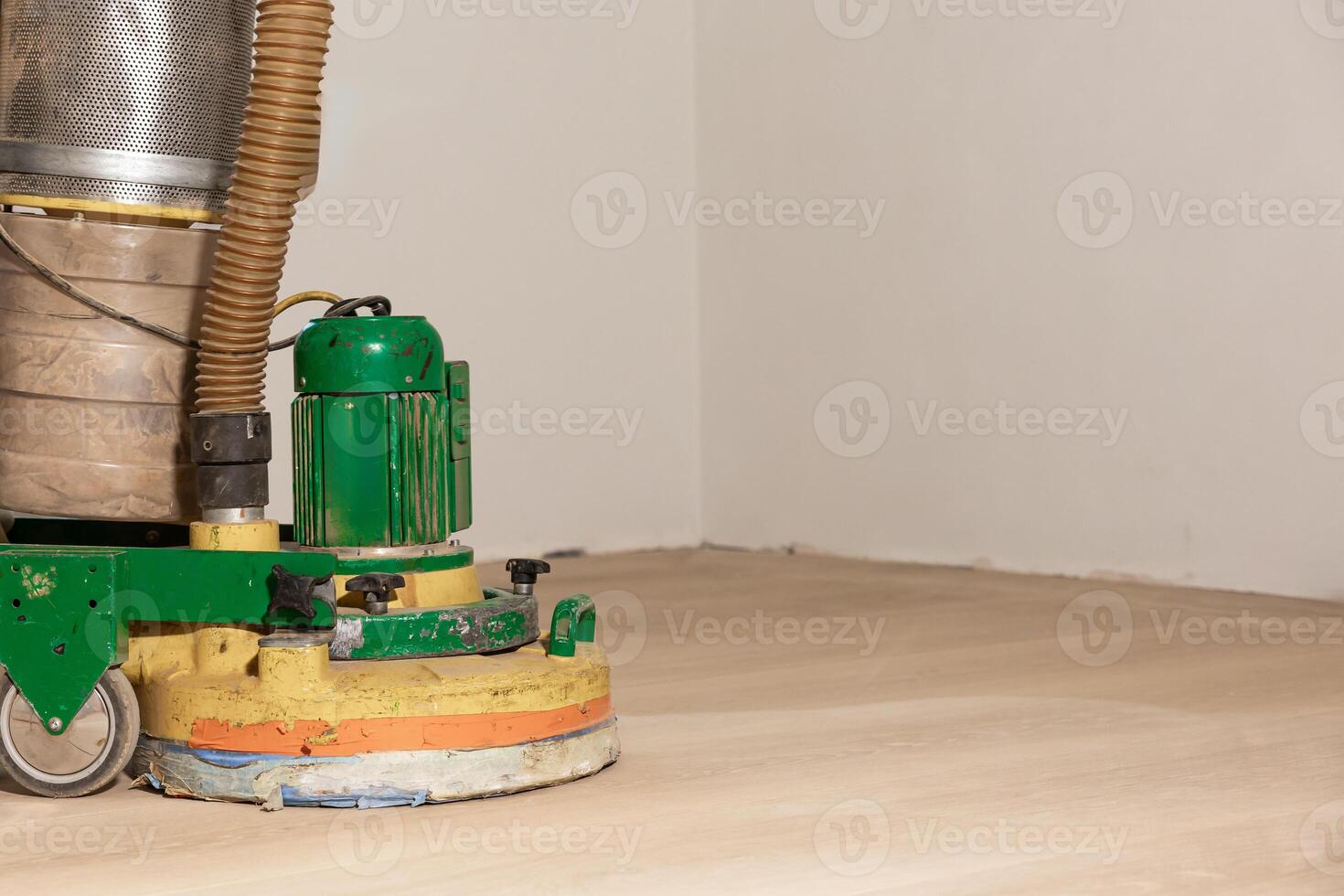 A green and yellow floor polishing machine with a dust collection system on a partially sanded wooden floor against plain off white walls. photo