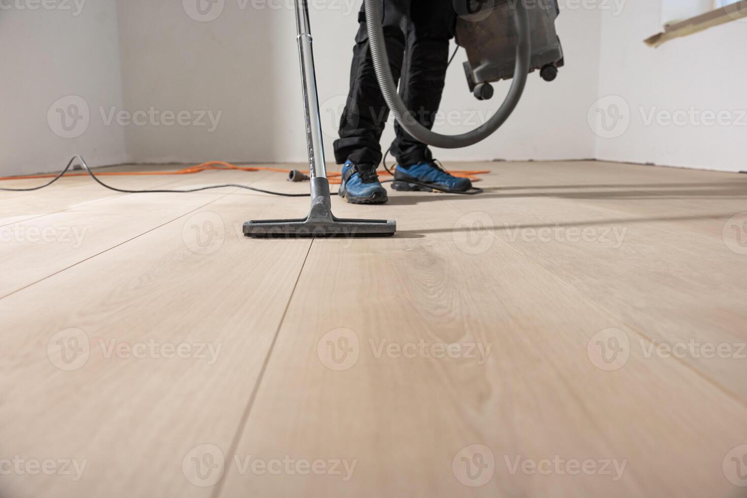 A worker operates a floor cleaning machine on a wooden floor in an empty room with white walls, a window, and an orange extension cord visible. photo