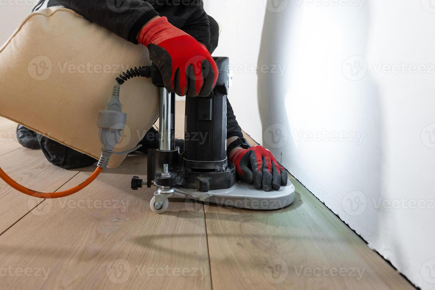 A worker kneels near a white wall, operating a floor polishing machine on light colored wooden flooring. An orange power cord connects to the machine. photo