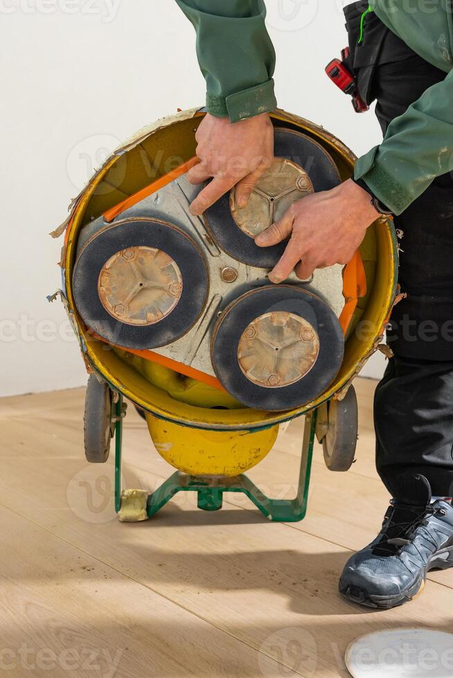 Individual in green jacket and black pants inspects a floor polishing machine with three circular pads on a light wooden floor near a white wall. photo