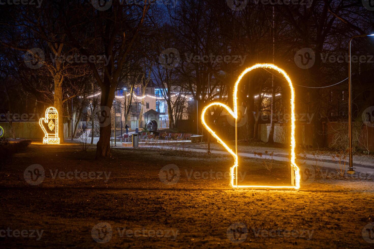 Illuminated mitten shaped lights line a pathway in a park at night, surrounded by bare trees. A well lit building with large windows is visible in the background. photo