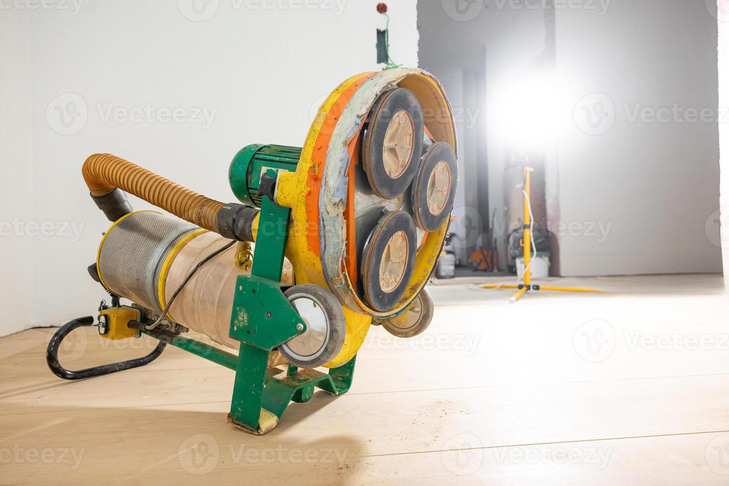 A yellow and green floor polishing machine with visible pads rests on a wooden floor. Bright light reveals white walls, tools, and an open doorway. photo