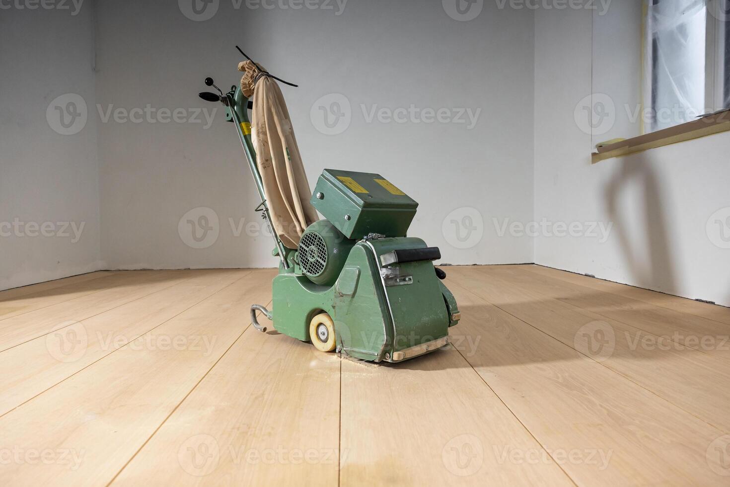 A green floor polishing machine sits on a light wooden floor in a minimalistic room with white walls and a window partially covered for maintenance. photo