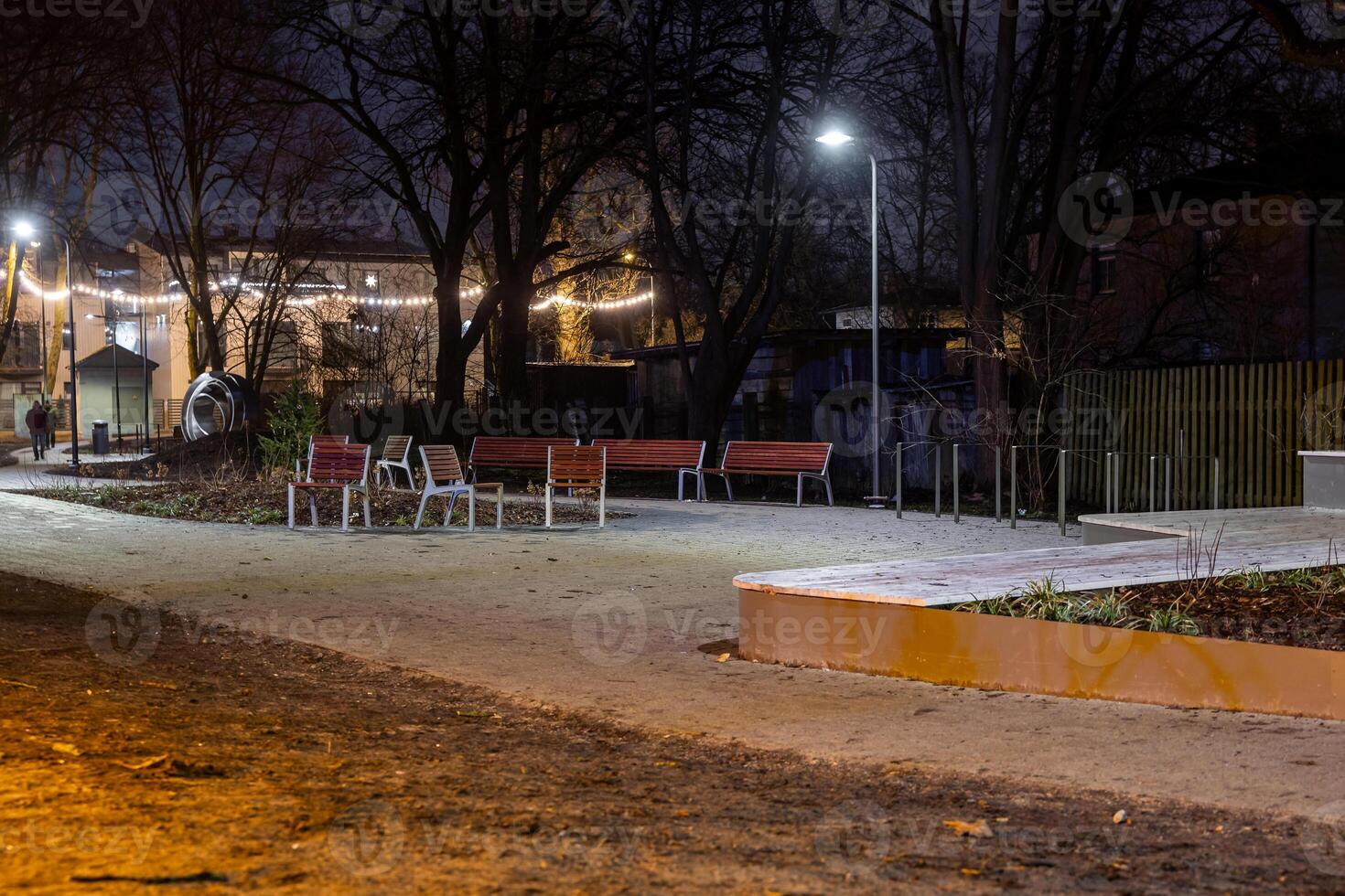 A night park scene with red benches, white chairs, string lights, bare trees, a paved pathway, a streetlamp, and a small structure in the distance. photo