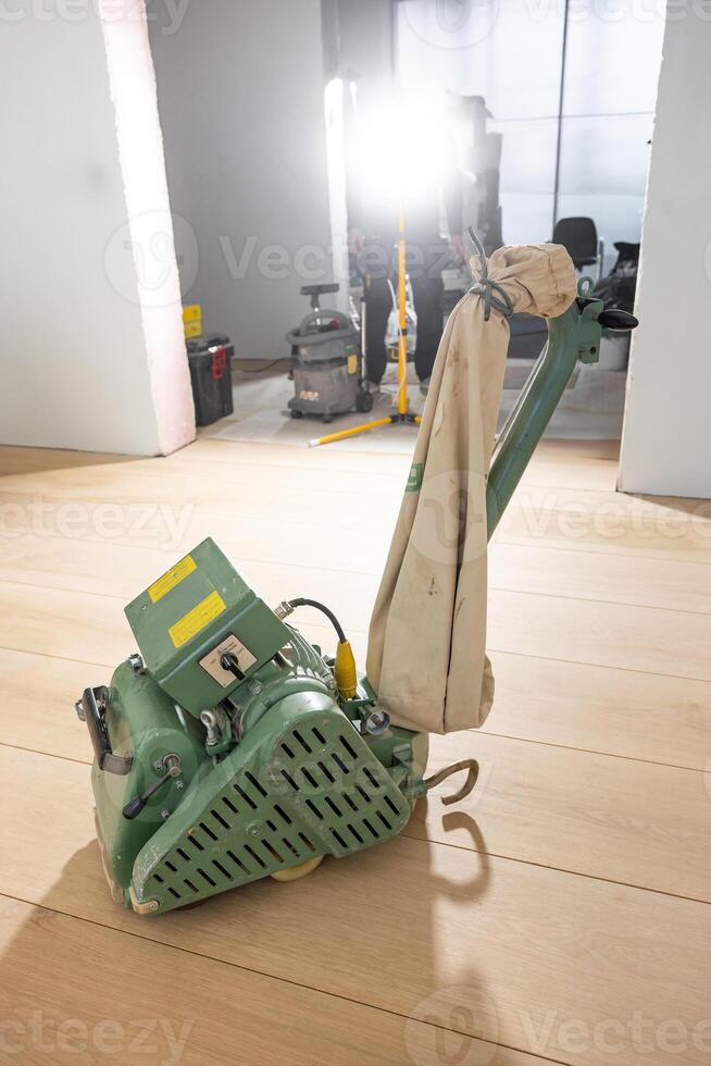 A green floor polishing machine with a beige dust bag on a light wooden floor. Construction tools and a work light are visible in the background. photo