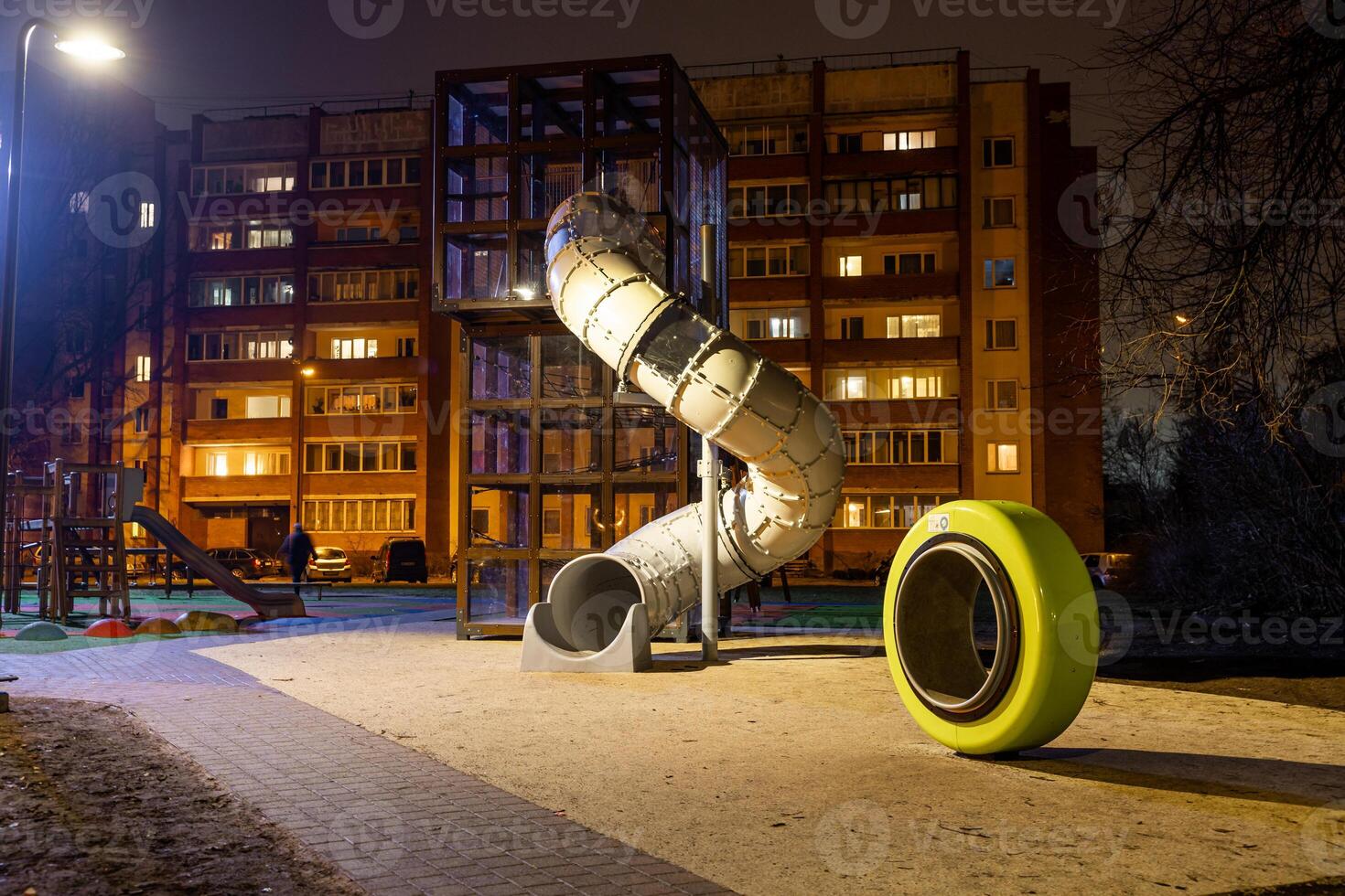 Nighttime view of a playground with a tall spiral slide, yellow play element, paved walkway, and a lit residential building in the background. photo