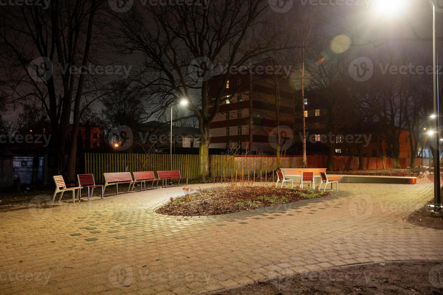 A small urban park at night with a curved pathway, red and white benches, sparse vegetation, and multi story buildings in the background. photo