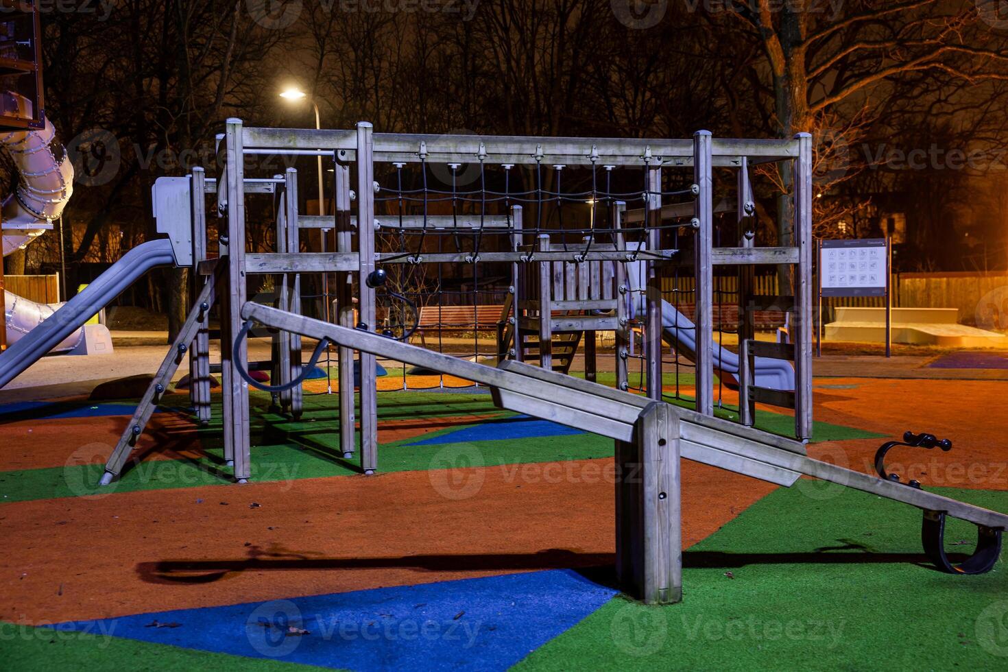 Nighttime view of a playground featuring a wooden play structure, slides, climbing nets, and seesaw. Colorful flooring and bare trees are visible. photo