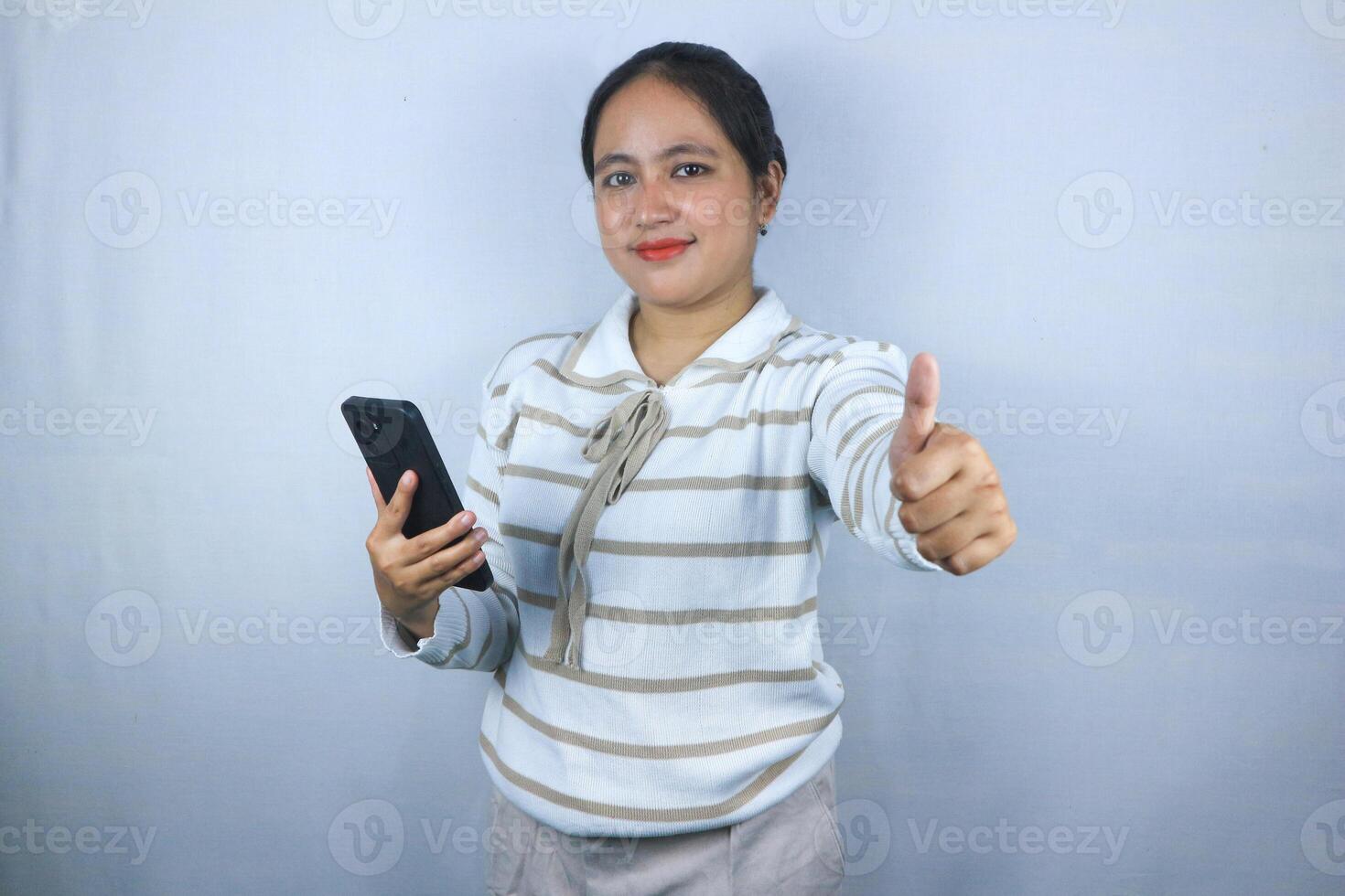 Beautiful smiling Asian woman in sweater showing thumbs up gesture with hand while holding phone photo