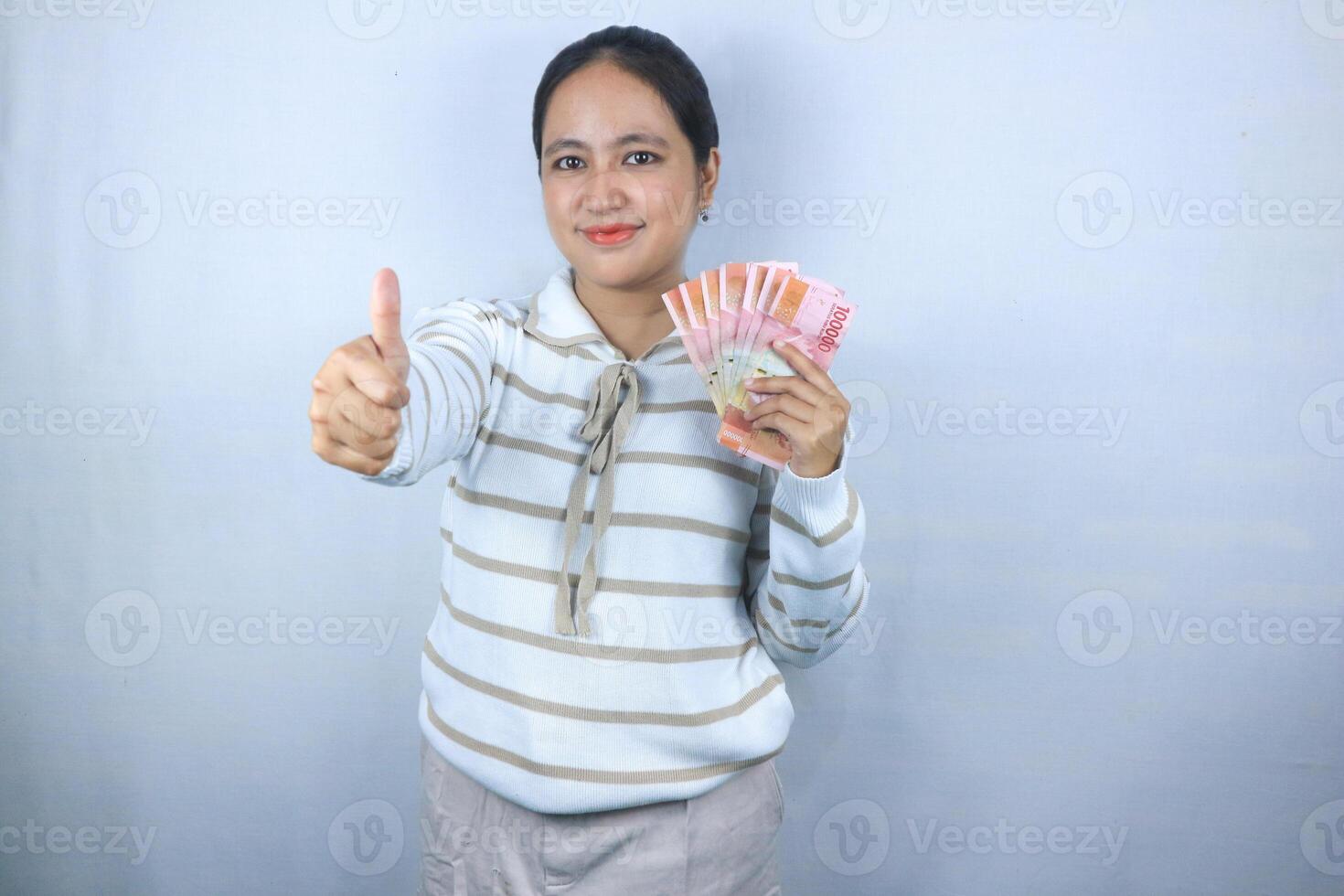 Beautiful asian woman in sweater hold indonesian banknote giving thumps up gesture photo