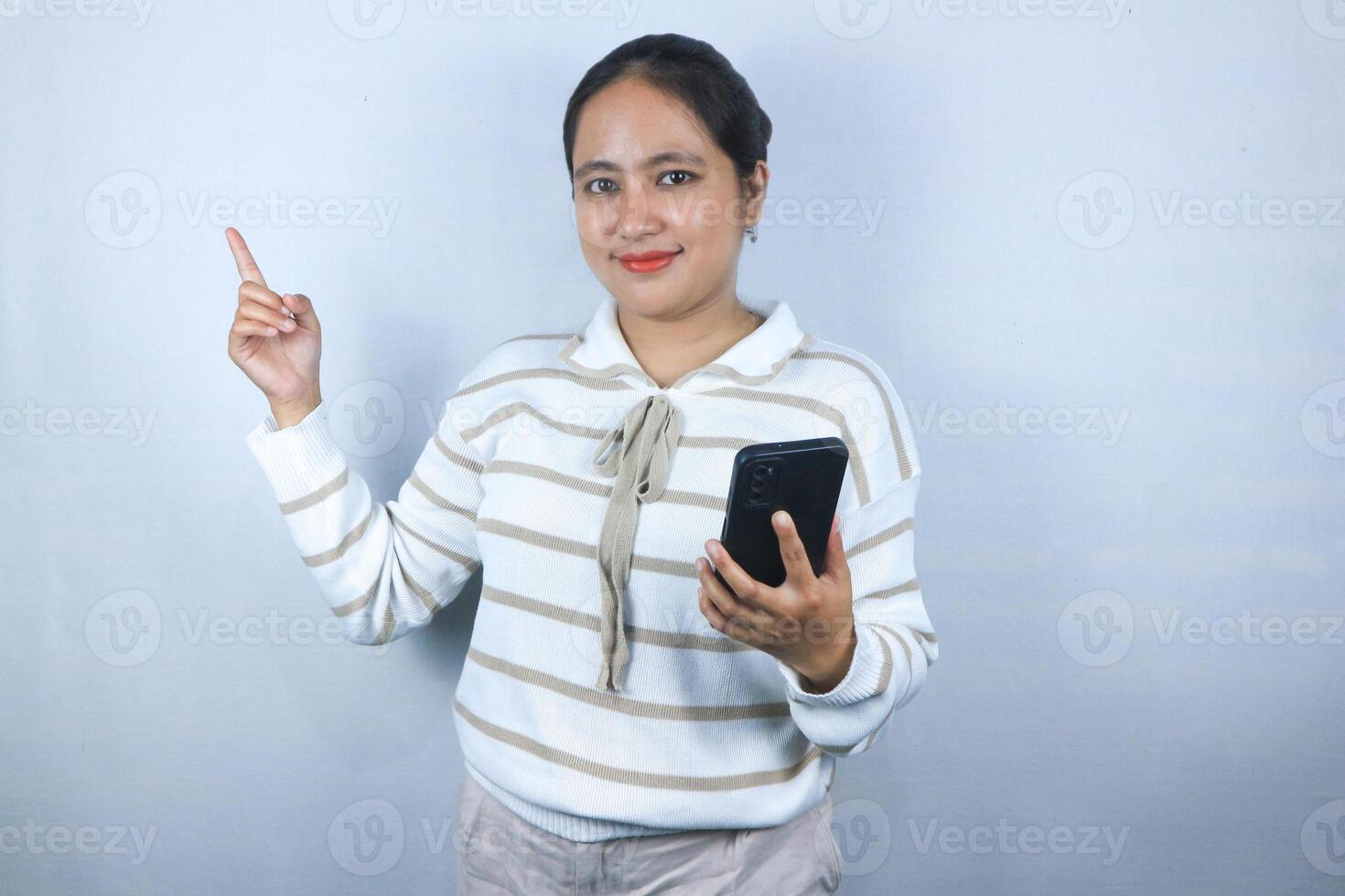 Beautiful smiling Asian woman in sweater pointing side with finger while holding phone photo