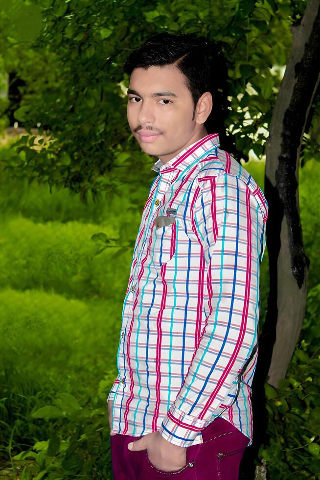 Portrait of a young man wearing a casual shirt and jeans, standing in a lush park with a relaxed demeanor photo