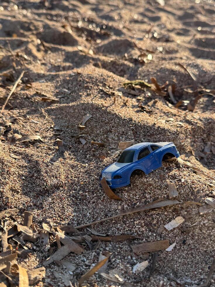 A blue toy car partially buried in sand on a beach, surrounded by textured sand and small twigs under warm sunlight. photo