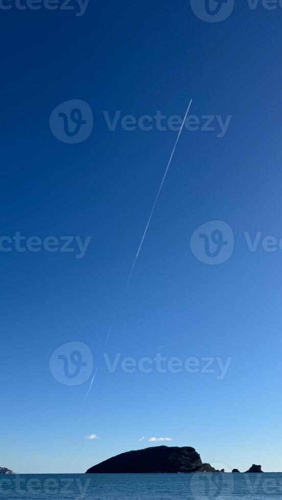 A clear blue sky with a long contrail stretching above a small island on calm sea waters . copy space photo