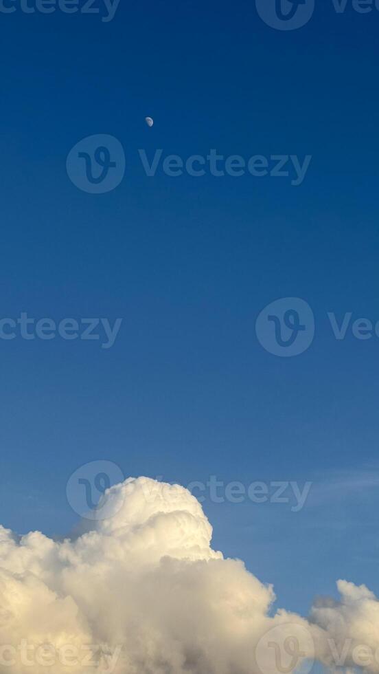 Minimalistic composition of crescent moon in deep blue sky above fluffy white clouds illuminated by sunlight. photo