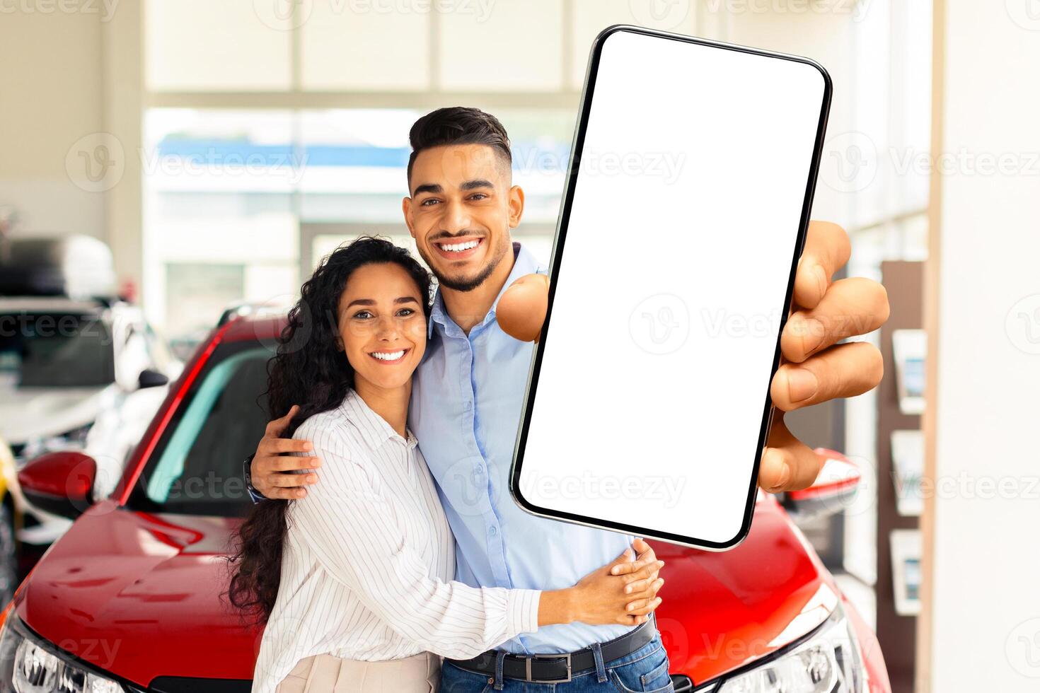 Positive young arabic couple in casual standing by brand new red car in showroom, showing cell phone with white empty screen, mockup, copy space. Auto dealership online concept photo