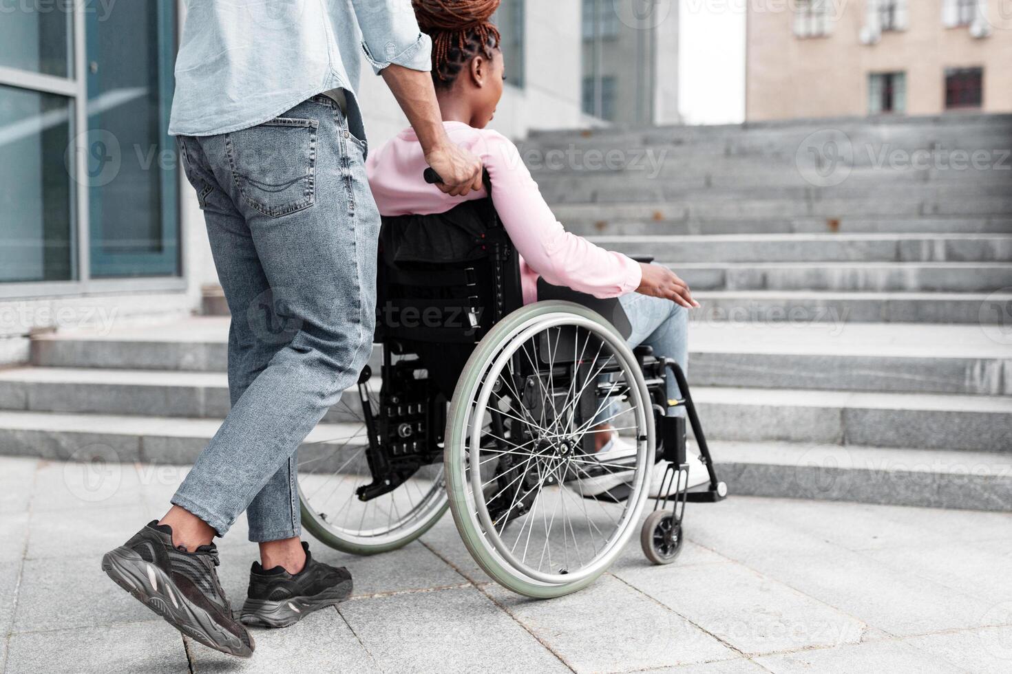 Young black guy pushing wheelchair with handicapped woman, having problem entering building, standing in front of stairs without ramp. Problems with accessible environment for disabled people photo