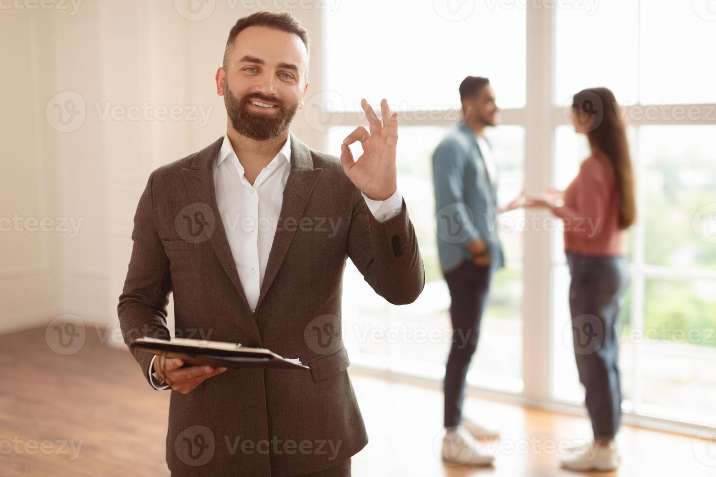 Successful Deal, House Ownership Concept. Portrait Of Smiling Professional Real Estate Agent In Suit Showing Okay Sign Gesture, Selective Focus. Buyers Talking In Blurred Background photo