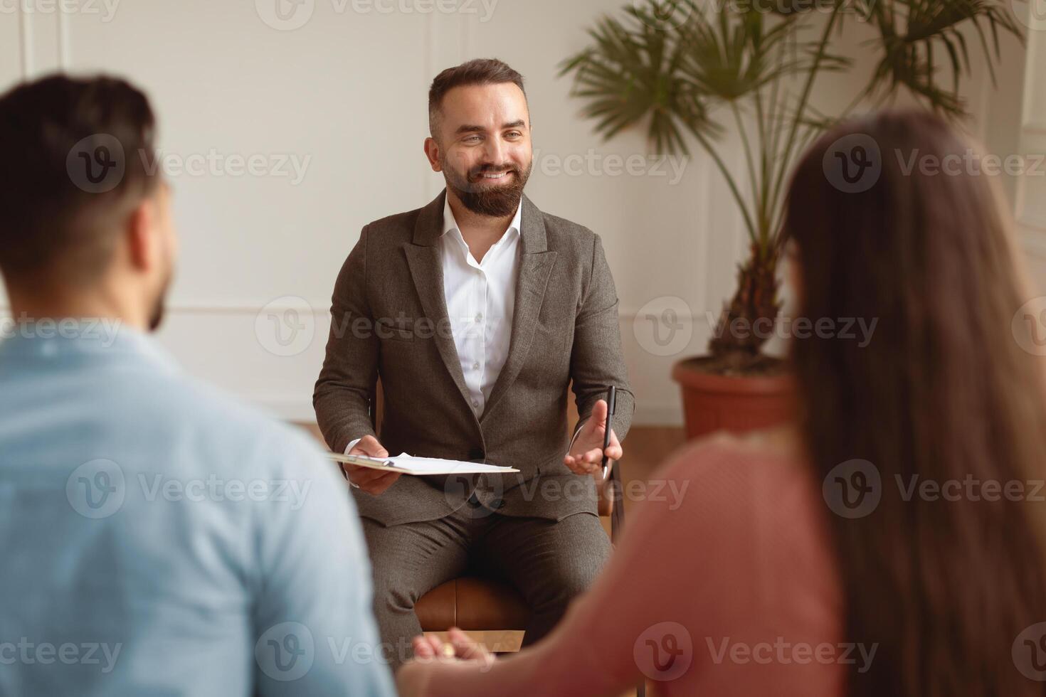 Smiling therapist talking with young millennial patients, back view of happy married couple spouses sitting on sofa holding hands visiting professional family counselor, resolving issues photo