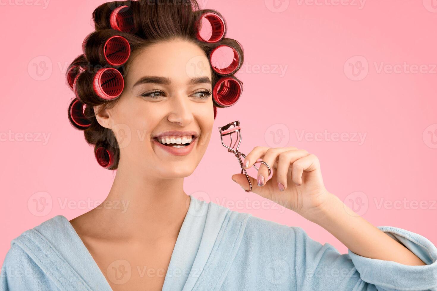 Cheerful Housewife Curling Eyelashes And Hair Using Hair Curlers Enjoying Beauty Routine Smiling Looking Aside Posing Over Yellow Studio Background. Makeup And Cosmetics Concept photo