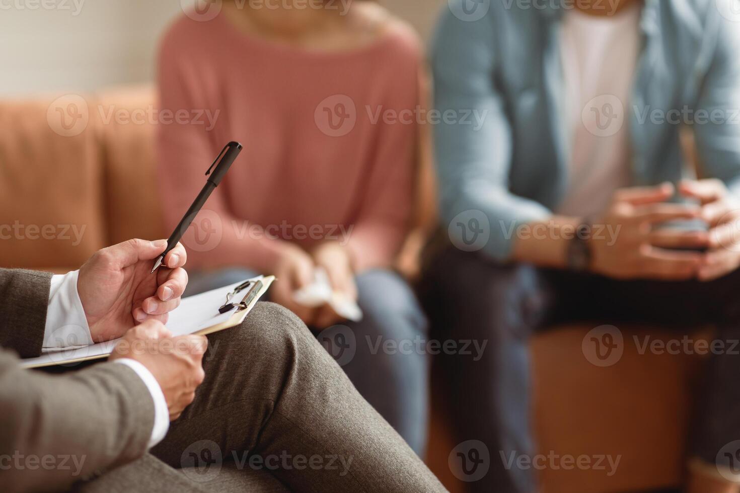 Unrecognizable couple having therapy session at psychotherapists office, selective focus on man professional psychologist holding pen and clipboard, writing in notepad, talking and consulting clients photo