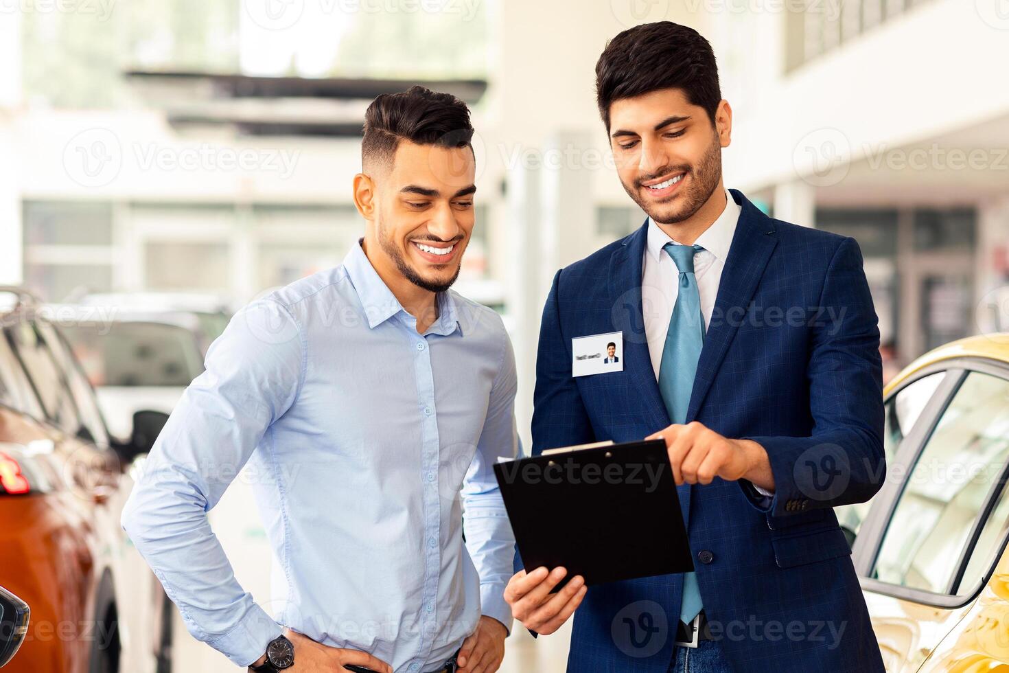 Handsome young arab guy having conversation with sales assistant in showroom, copy space. Cheerful car dealer showing catalogue with luxury autos, selling nice automobiles in new salon photo