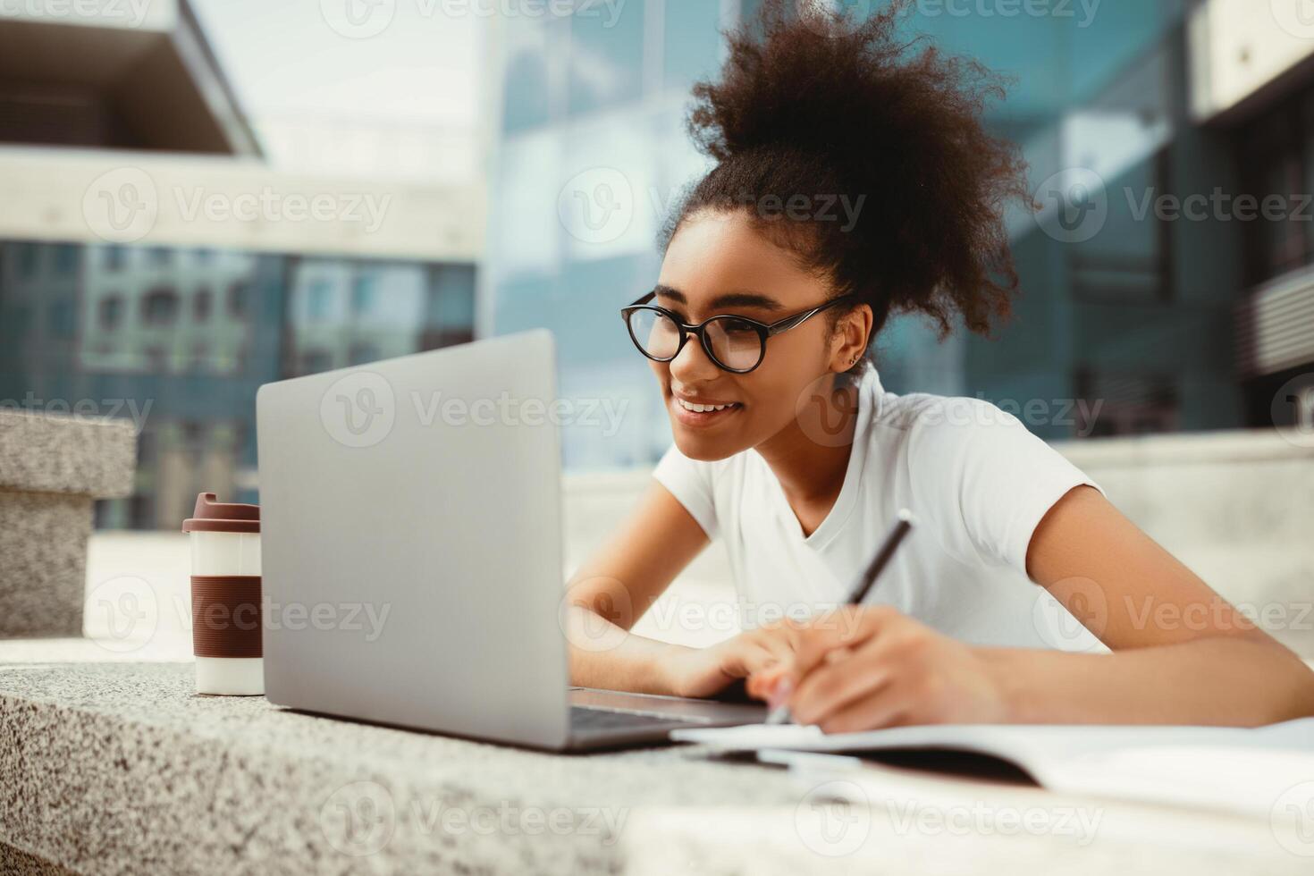 Happy Black Student Girl Doing Homework Using Laptop Taking Notes Watching Online Lecture Sitting Near Modern College Building Outdoor. Modern Remote Education, E-Learning And Studentship photo