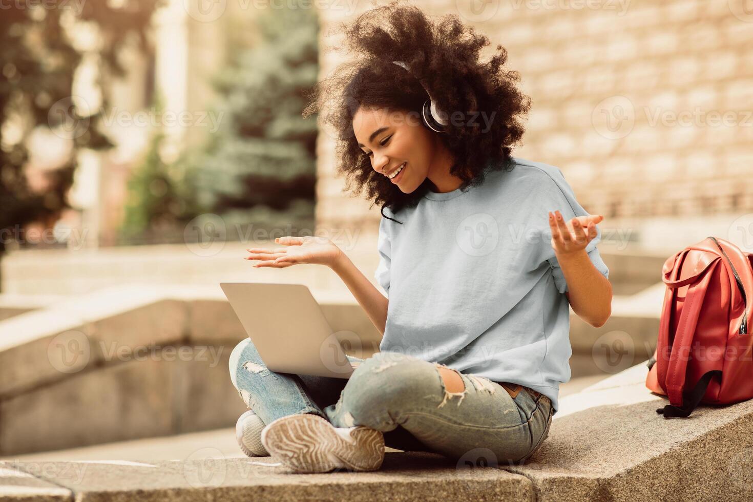African American Female Student Using Laptop Making Call During Remote Class Sitting Outside Of College Building. Distance Education And E-Learning. Modern Communication. Side View photo
