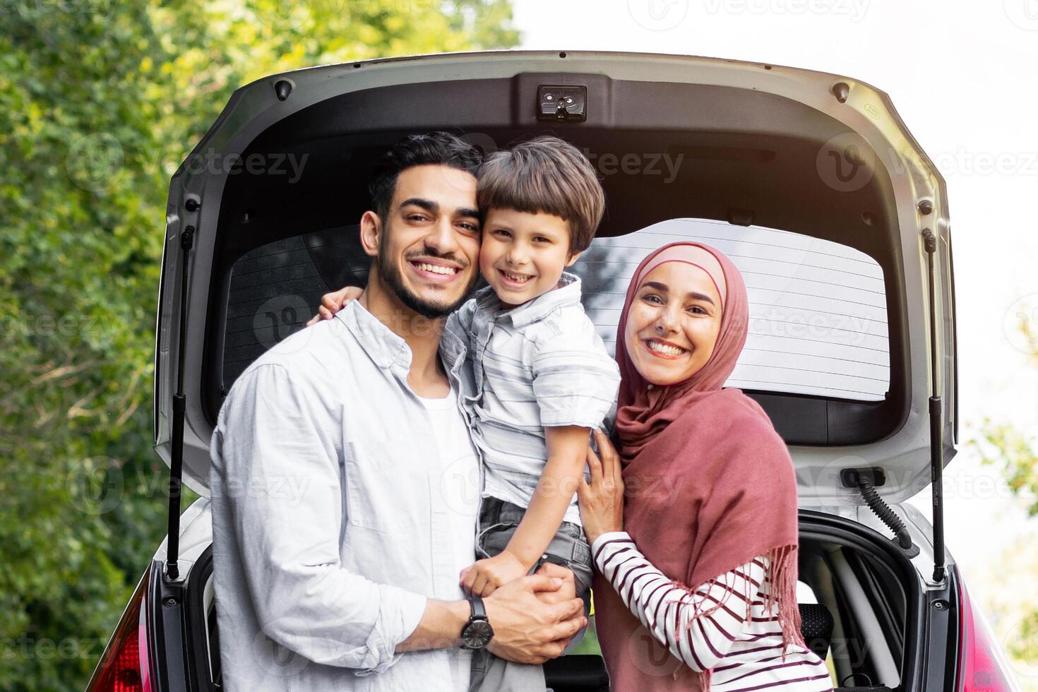 Rest outdoor from road, family journey at summer. Satisfied millennial arab man and lady in hijab holding baby near car with open trunk, copy space. Trip, travel together, vacation and people emotions photo