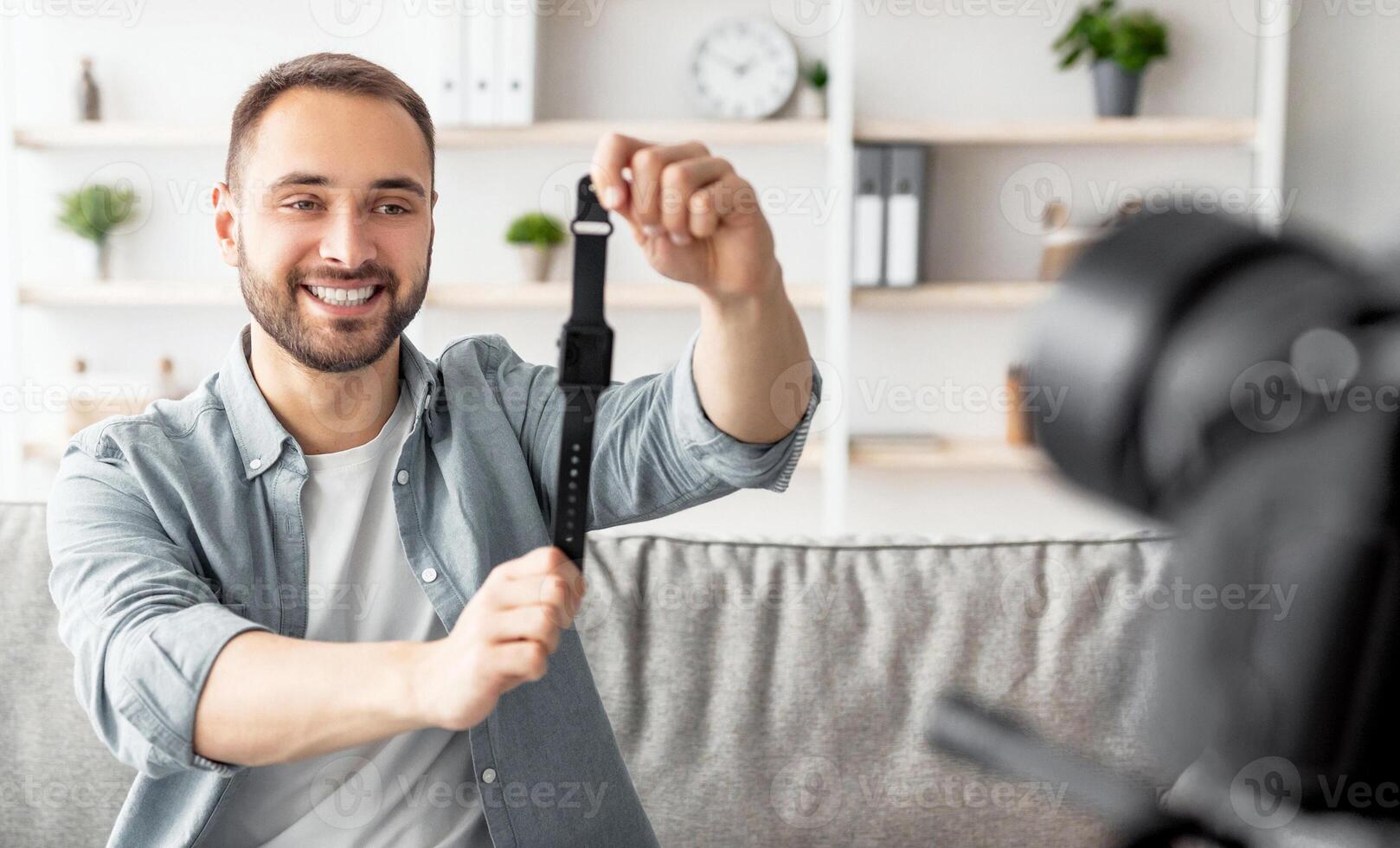 Friendly male blogger shooting review of new smartwatch model, using professional camera at home. Happy content maker promoting contemporary gadget, broadcasting for social network photo