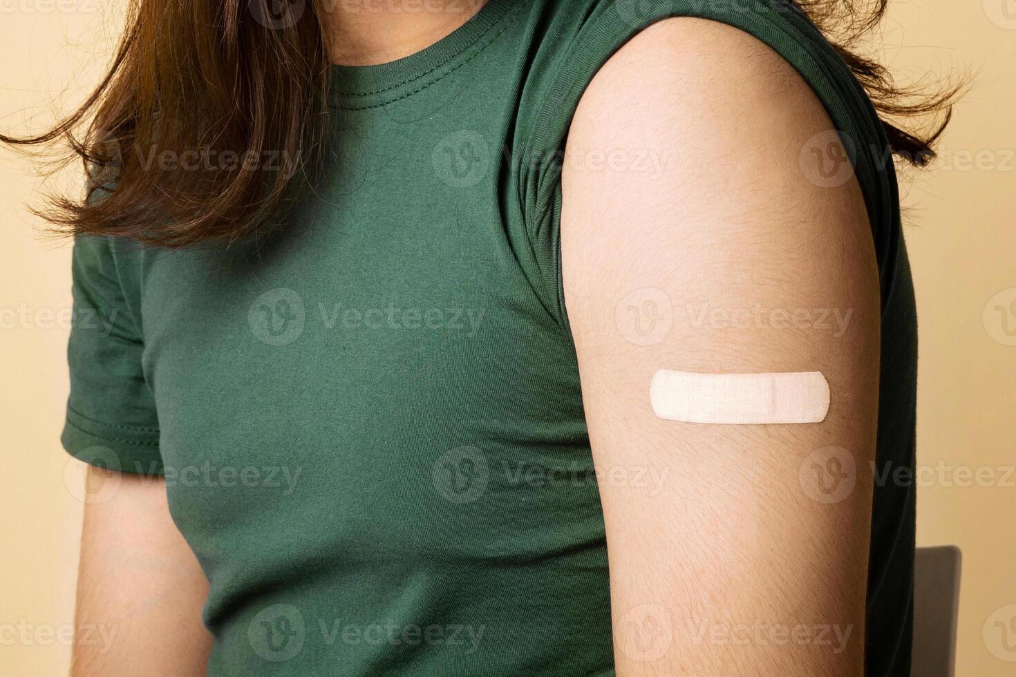 Millennial indian man in medical face mask showing adhesive band on his shoulder after vaccination against coronavirus, beige studio background, copy space. Mass immunization while pandemic concept photo